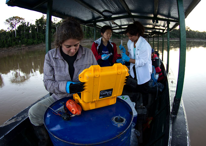 Three people are standing in a boat. Behind them are trees along the river bank. The person in the foreground is looking into a yellow box, on top of a blue steel barrel. Everyone is wearing blue gloves.